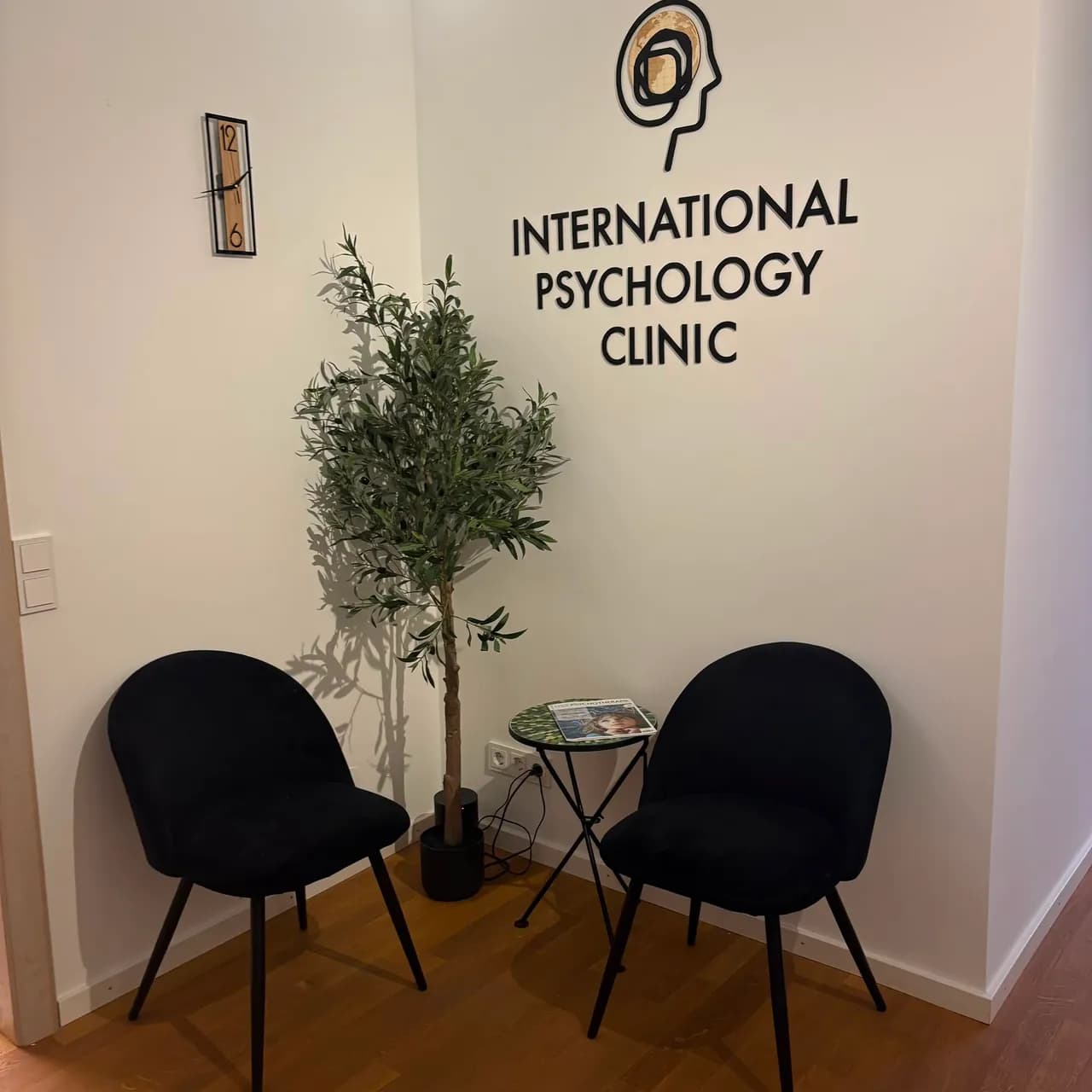 Empfangsecke mit dem Namen und Logo der International Psychology Clinic an der Wand, zwei schwarzen Sesseln und einem Olivenbaum