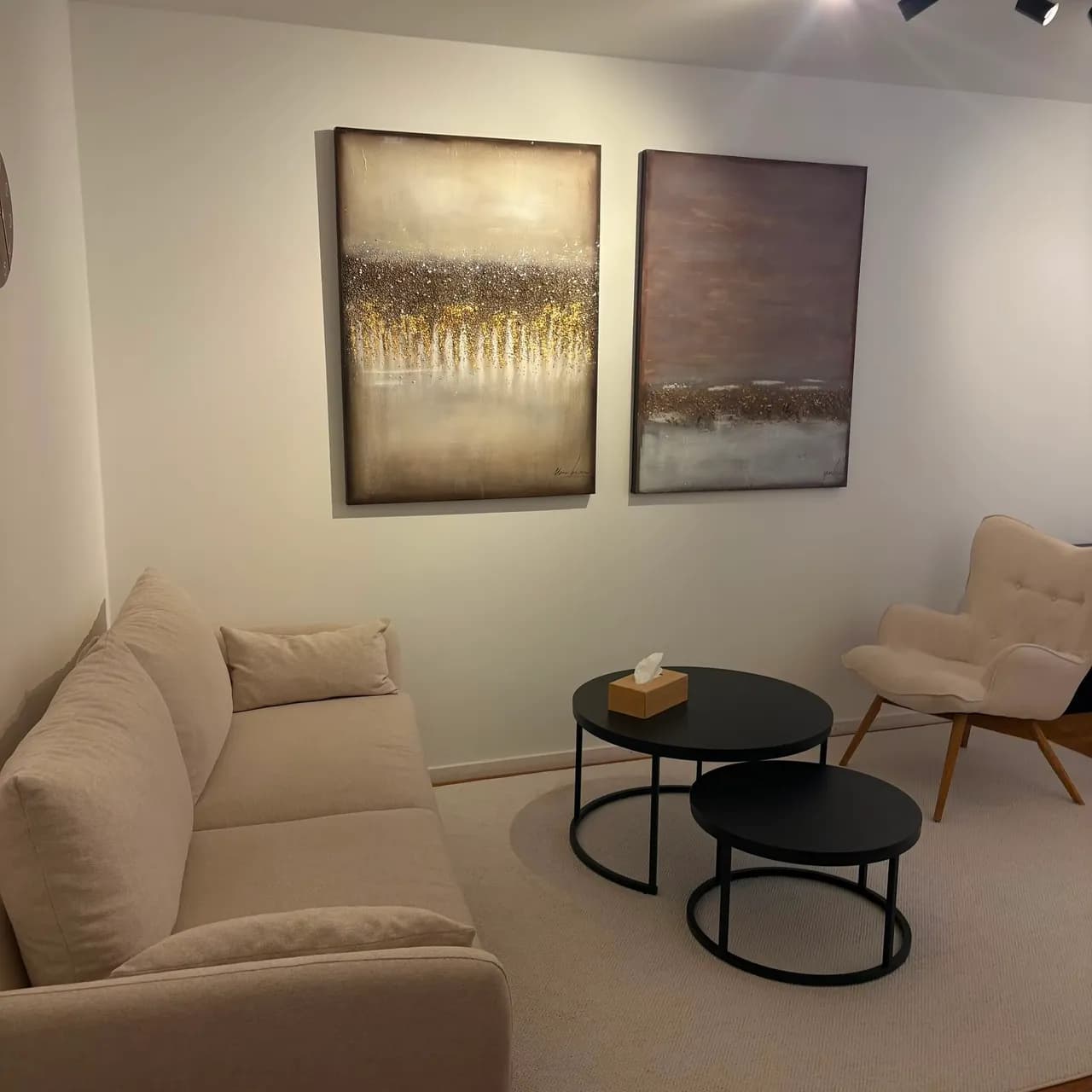 Therapieraum in warmen Beigetönen, mit Sofa, Sessel und runden Tischen unter zwei goldfarbenen abstrakten Bildern