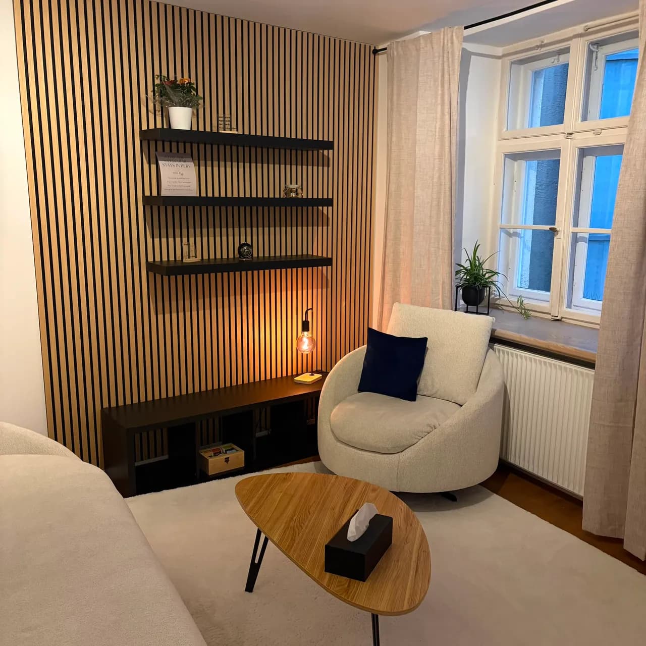 Hell gehaltener Therapieraum mit cremefarbenem Sessel und Sofa, einem Eichentisch und einer Holzlamellenwand am Fenster